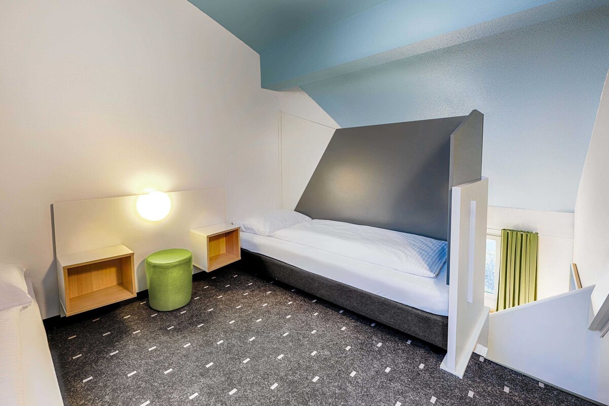 A modern bedroom with a bed, bedside tables, a green stool, and a wall-mounted lamp.
