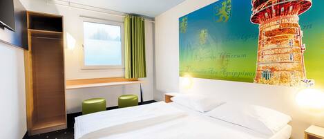 A hotel room with a bed, a window with curtains, a desk with a chair, and a wall with a decorative mural.