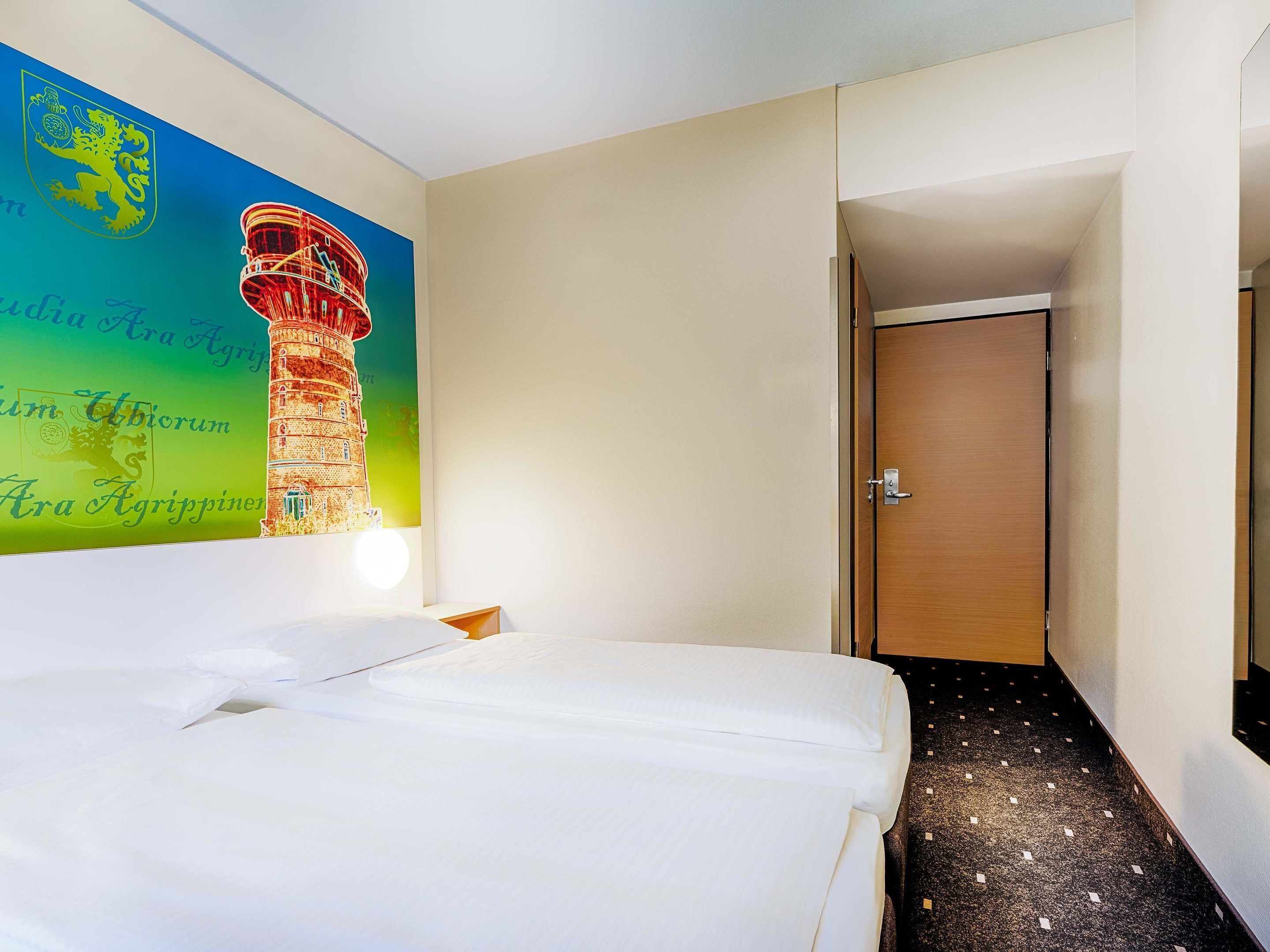 A hotel room with a bed, a wall with a colorful mural, a wooden door, and a carpeted floor.