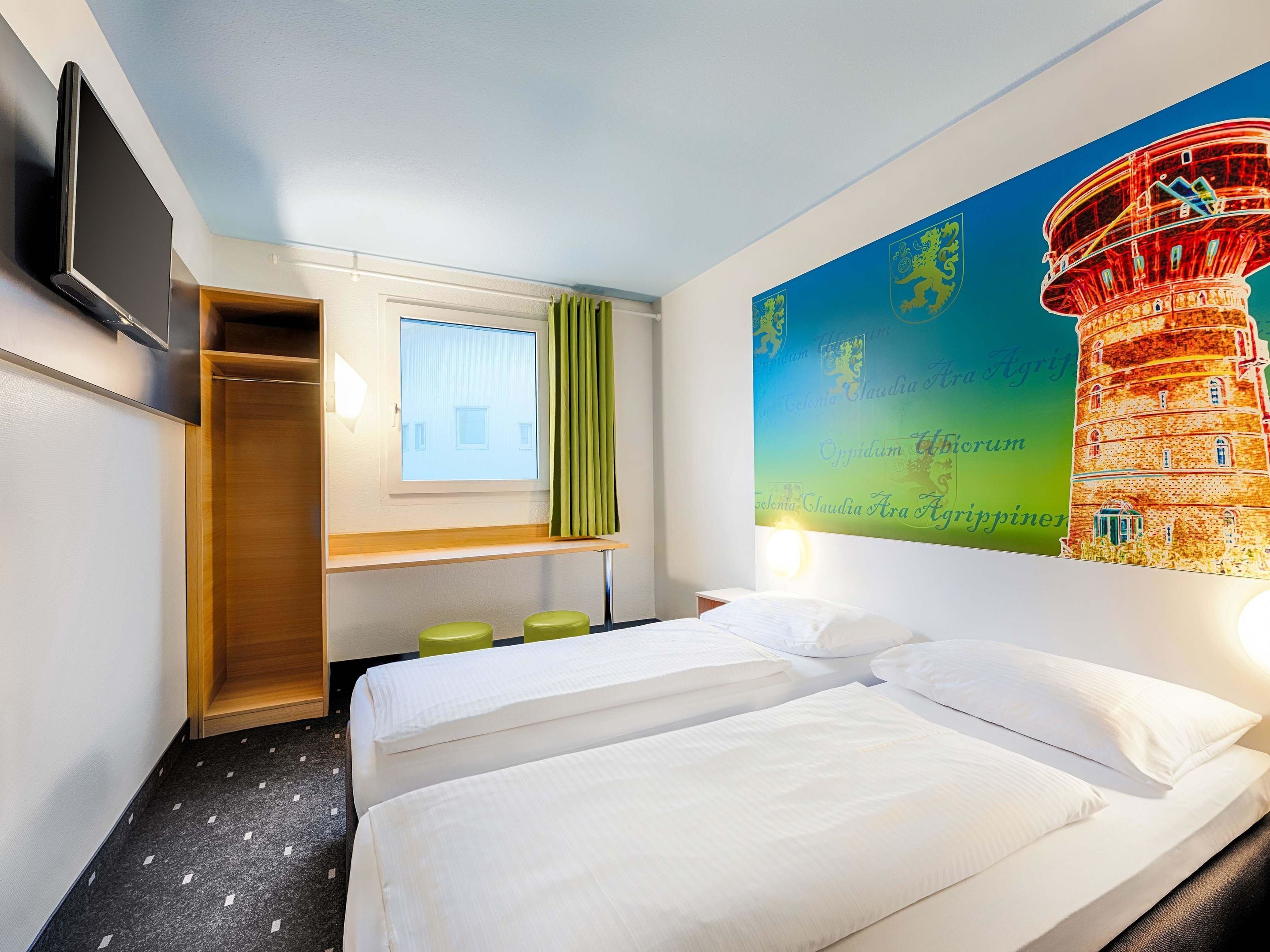 A hotel room with two beds, a TV, a window with curtains, a desk, and a wall with a colorful mural.