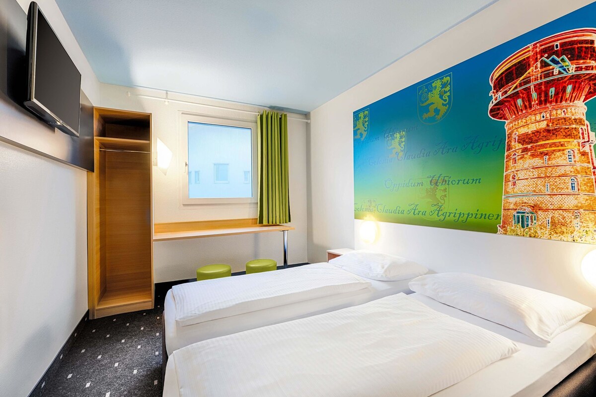 A hotel room with two beds, a TV, a window with curtains, a desk, and a wall with a colorful mural.