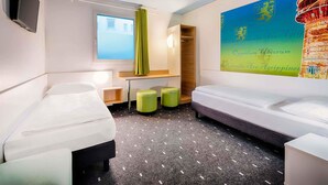Una habitación de hotel con cama, una mesita, dos taburetes verdes, un televisor montado en la pared y un mural grande y colorido.