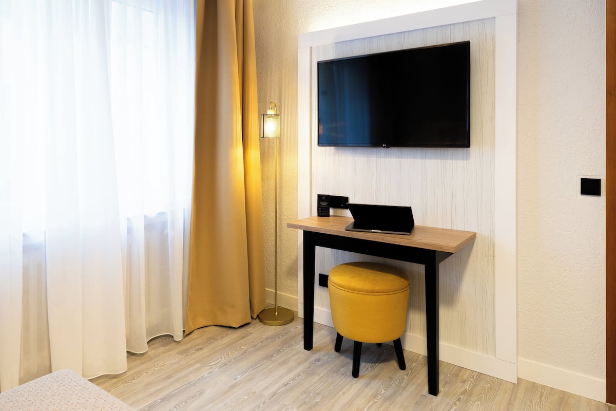 Ein modernes Hotelzimmer mit Holzboden, einem an der Wand befestigten Flachbildfernseher, einem kleinen Holztisch mit einem Laptop, einem gelben Hocker und einem großen Fenster mit durchsichtigen Vorhängen.