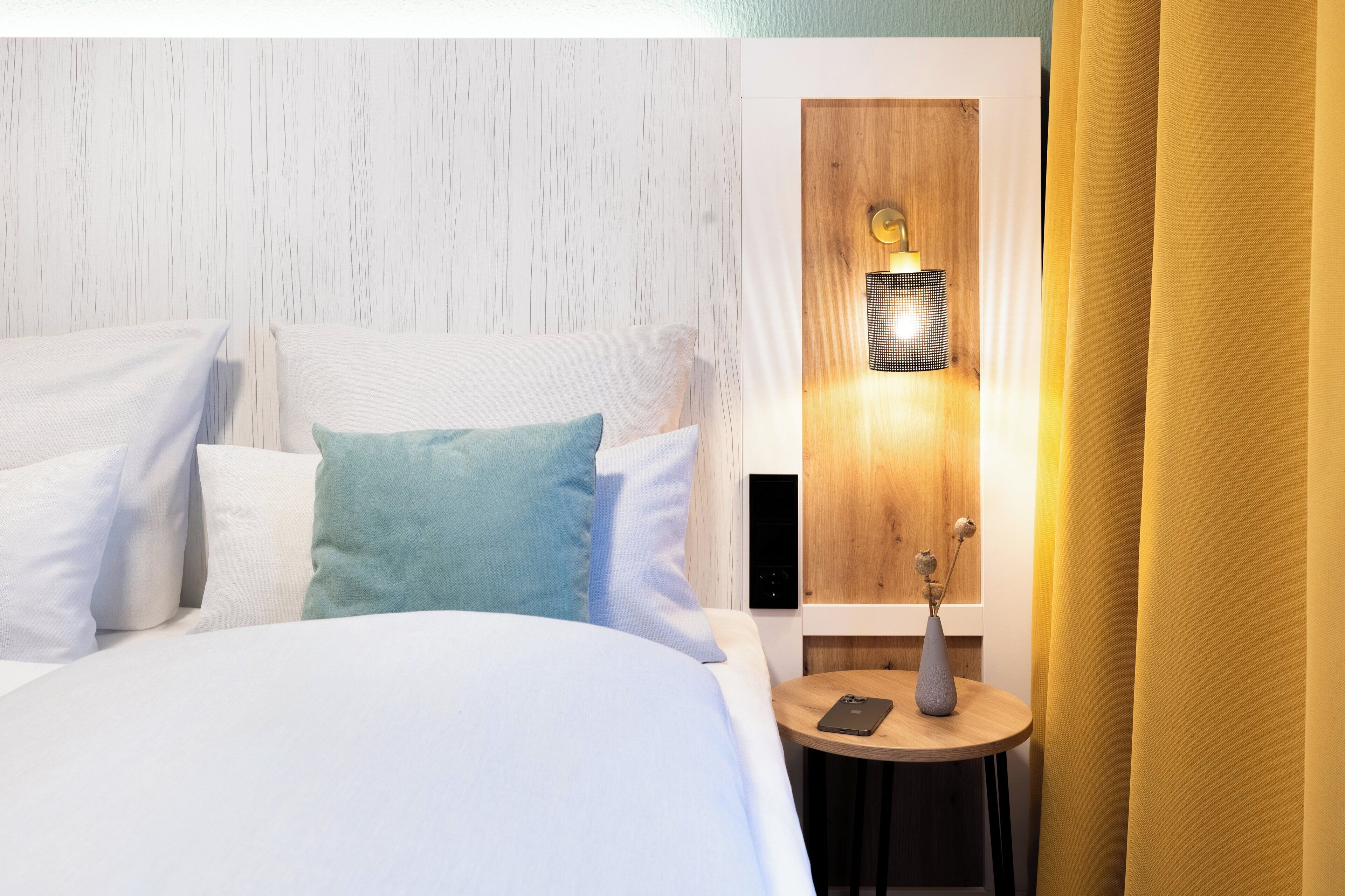 A neatly made bed with white and light blue pillows, a small round table with a vase and a phone, and a wall-mounted lamp.