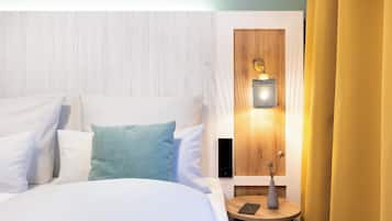 A neatly made bed with white and light blue pillows, a small round table with a vase and a phone, and a wall-mounted lamp.