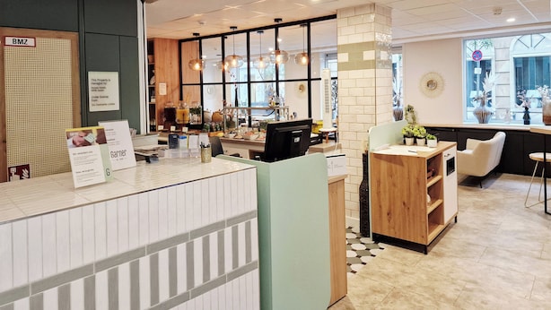 A reception area with a counter, a wooden cabinet, a white chair, and a window with a view. - Garner Hotel Cologne Engelbertz by IHG (Cologne)