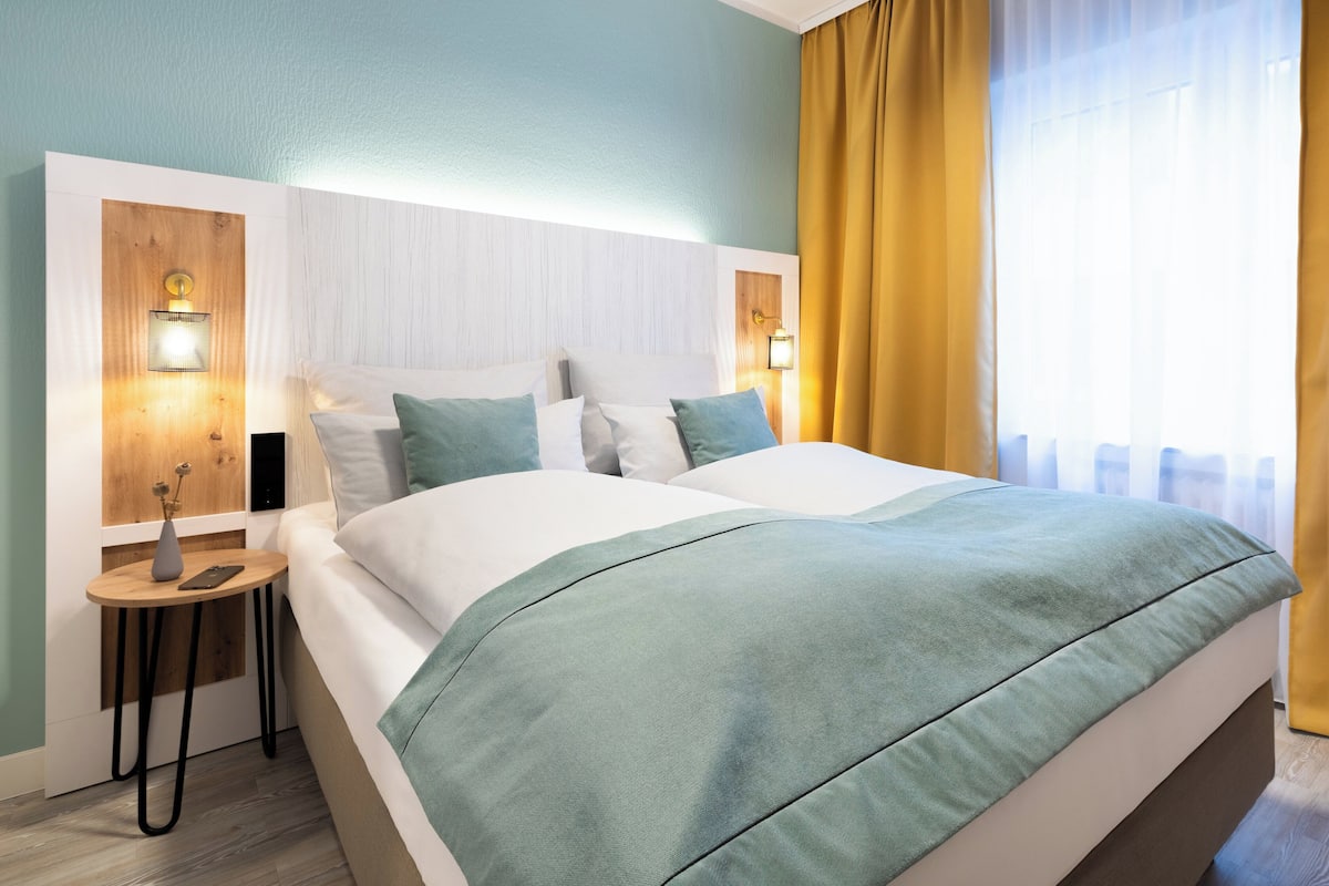 Ein modernes Hotelzimmer mit einem großen Bett, zwei Nachttischen mit Lampen und Blick ins Freie durch ein Fenster mit Vorhängen.
