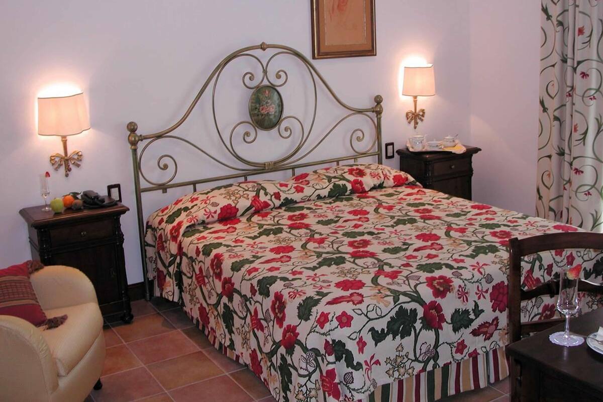 Une chambre à coucher avec une couvre-lit à motifs floraux, deux lampes de chevet, un tableau encadré au mur et une chaise dans un coin.