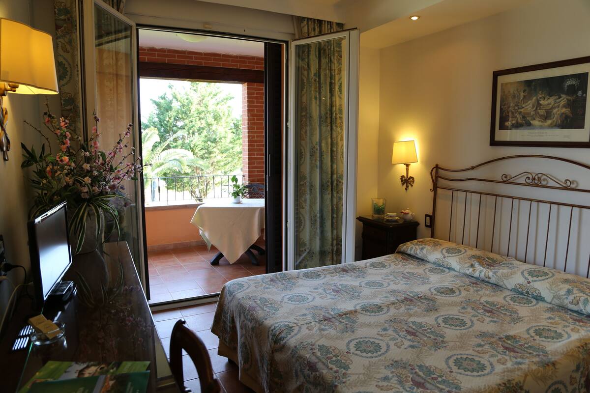 Une chambre d’hôtel comprenant un lit, un bureau avec un ordinateur, une chaise, une table avec une plante et un balcon avec une table et des chaises.