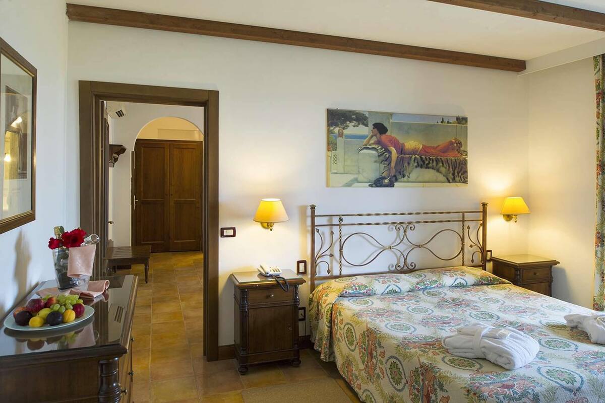 Une chambre d’hôtel avec un lit, deux tables de chevet, une petite table, un bureau agrémenté d’un vase de fleurs et un tableau au mur.