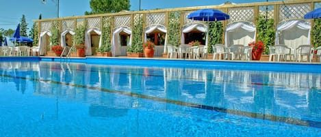 Una piscina con sillas de descanso y sombrillas, rodeada por una valla de madera con cortinas blancas.