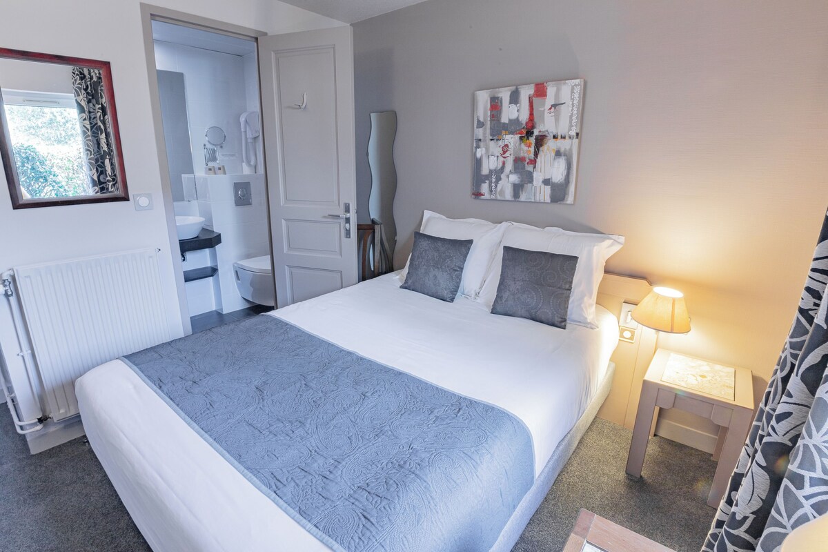 A neatly arranged hotel room with a large bed, bedside table with a lamp, a bathroom with a toilet and sink, and a painting on the wall.