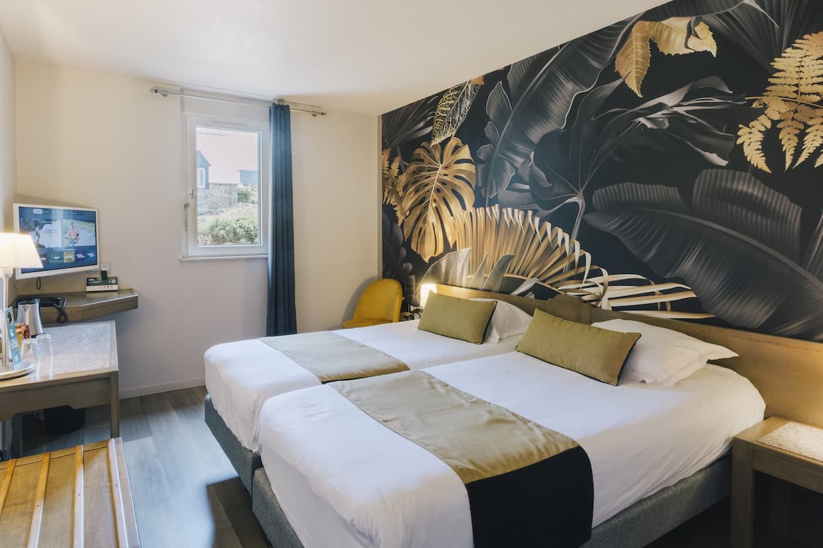 A hotel room with two beds, a large tropical leaf mural, a TV, and a small table with a lamp.