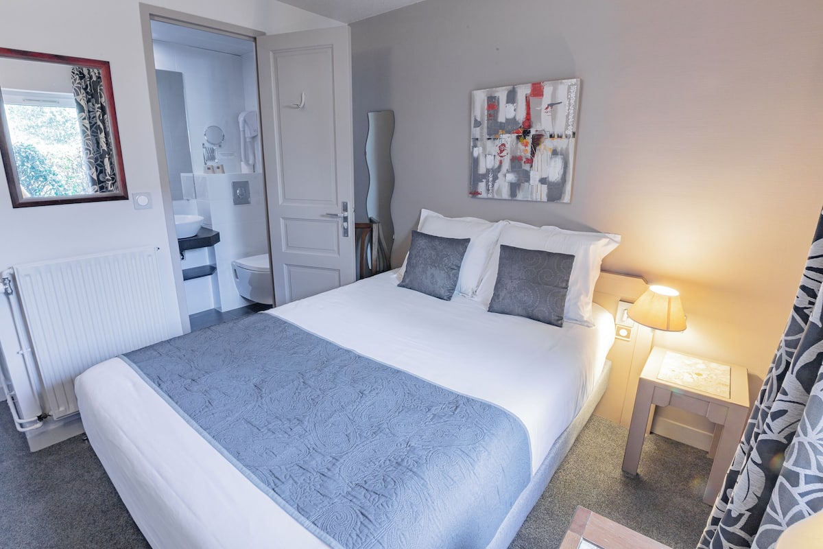 A neatly arranged hotel room with a large bed, bedside table with a lamp, a bathroom with a toilet and sink, and a painting on the wall.