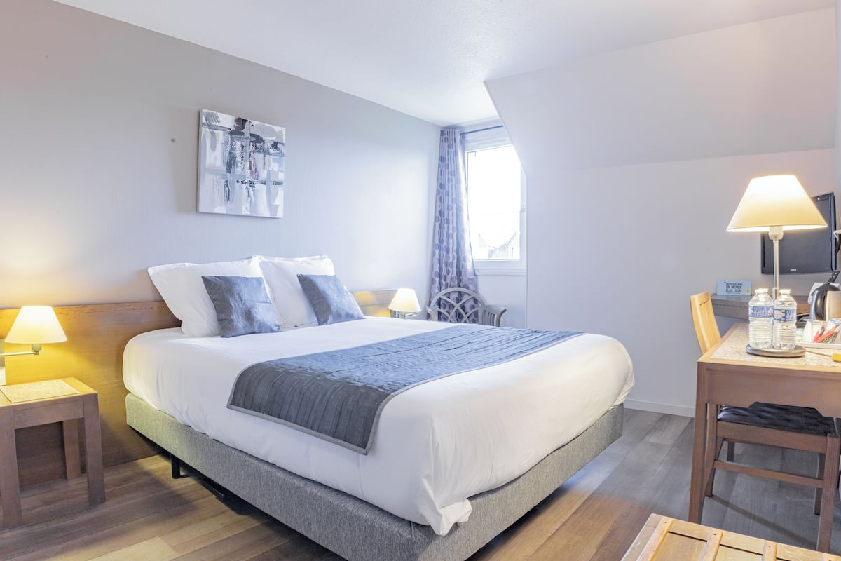 A neatly arranged hotel room with a large bed, a bedside table with a lamp, a chair, and a desk with a lamp and a bottle.