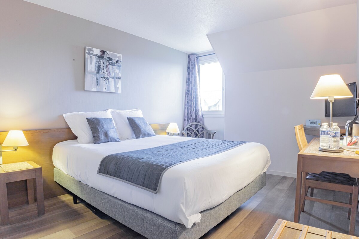 A neatly arranged hotel room with a large bed, a bedside table with a lamp, a chair, and a desk with a lamp and a bottle.