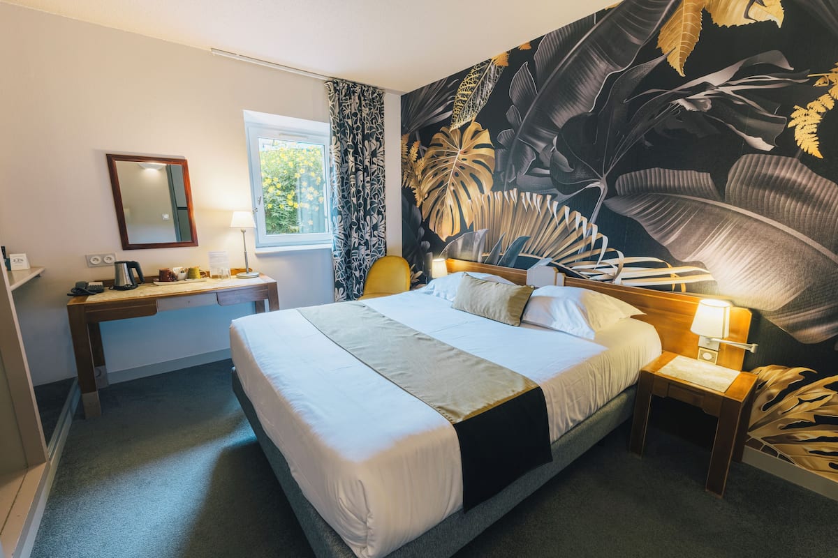 A hotel room with a bed, a desk, a chair, a nightstand, a lamp, a window, and a large tropical leaf mural on the wall.