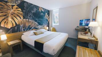 A hotel room with a large bed, a desk with a computer, a TV, and a mural of tropical leaves on the wall.