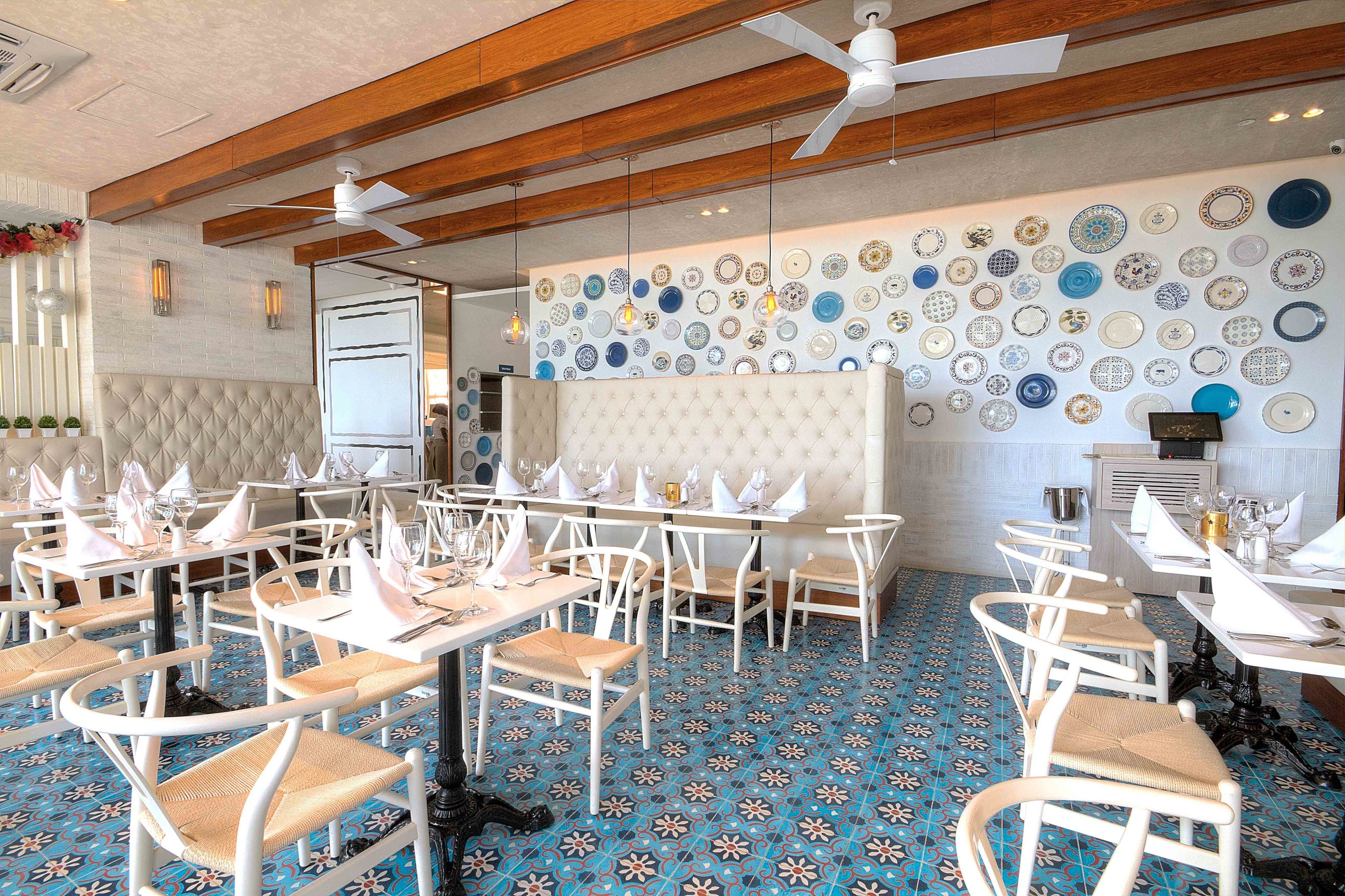 Un restaurante con mesas y sillas blancas, piso estampado y una pared decorada con platos.
