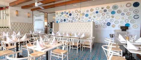 Un restaurante con mesas y sillas blancas, piso estampado y una pared decorada con platos.