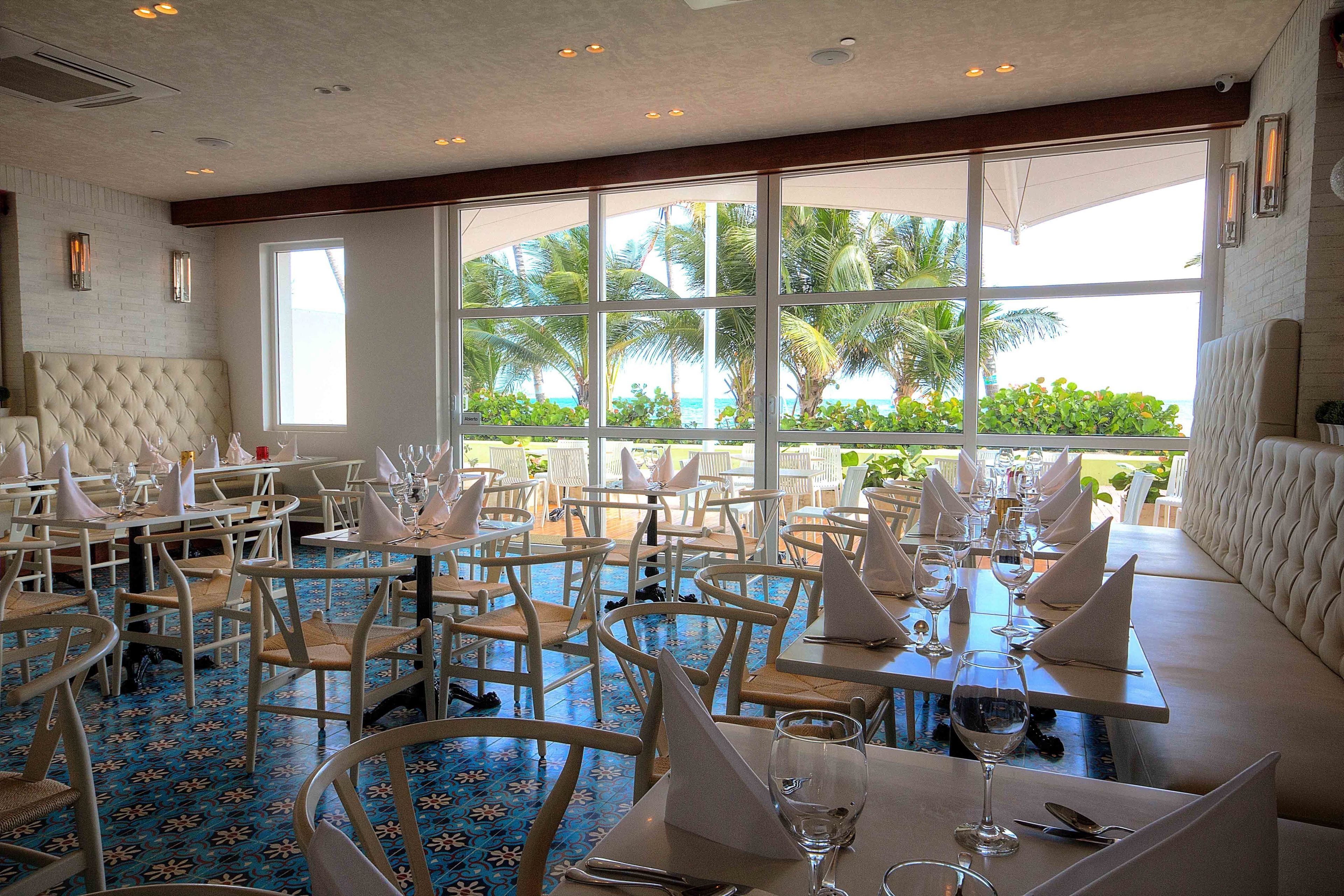 Un restaurante con asientos blancos acolchados, mesas dispuestas con copas de vino y amplias ventanas que ofrecen vistas a palmeras y al océano.