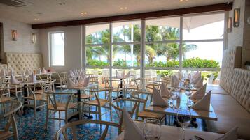 Un restaurante con asientos blancos acolchados, mesas dispuestas con copas de vino y amplias ventanas que ofrecen vistas a palmeras y al océano.