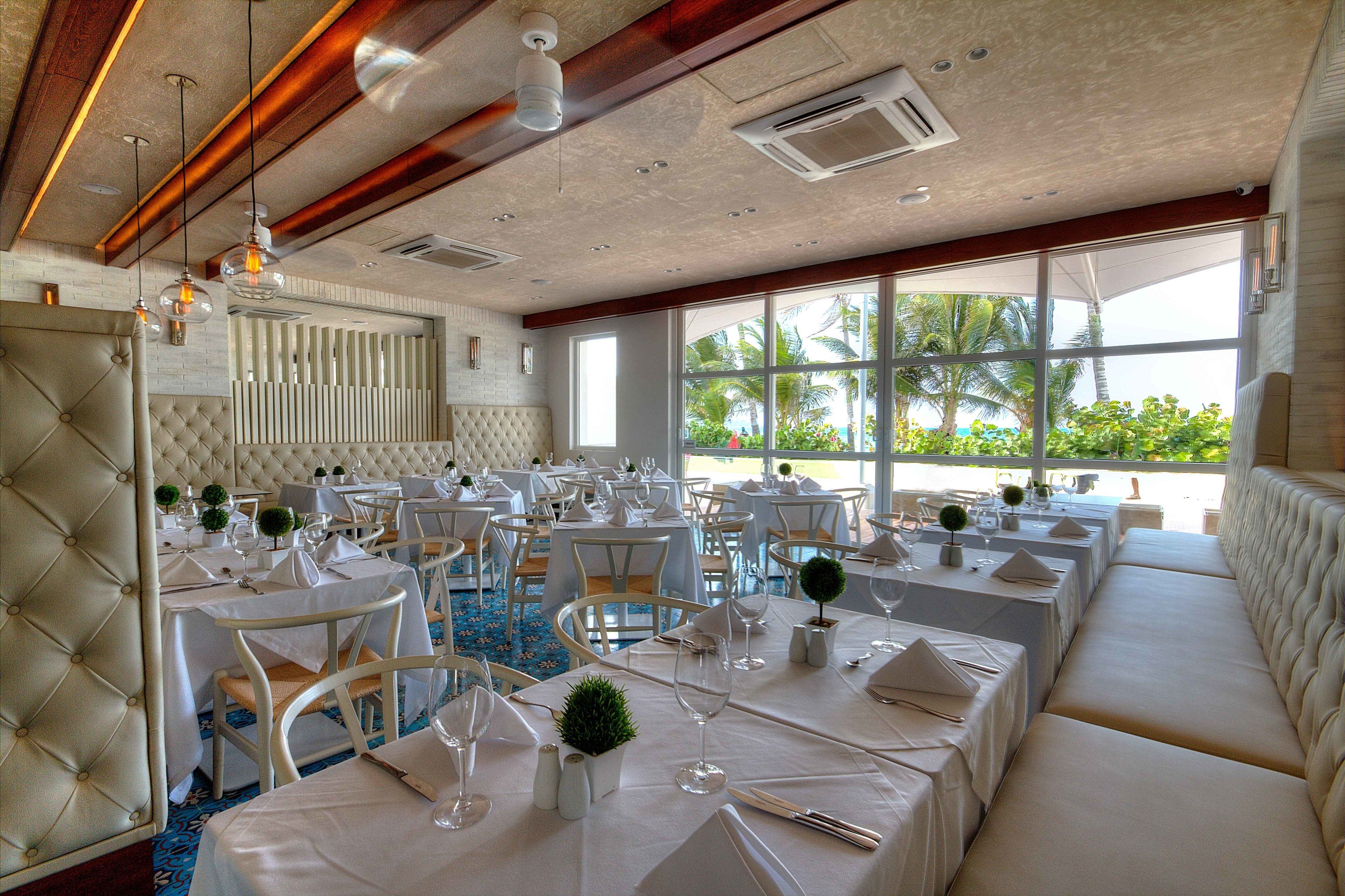 A spacious dining area with multiple tables set for service, large windows offering a view of palm trees, and a ceiling with exposed beams and recessed lighting.