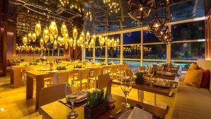 A restaurant with multiple dining tables set for guests, large windows overlooking a pool, and hanging decorative lights.