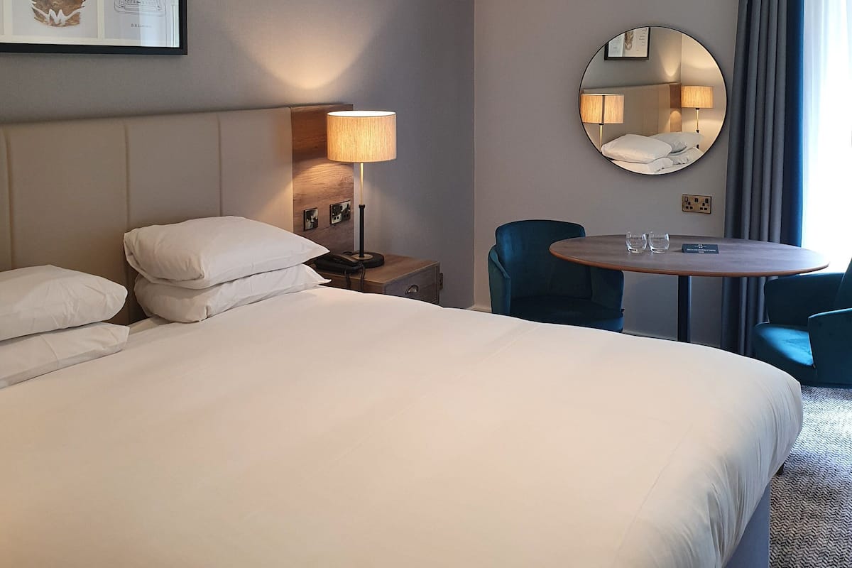A hotel room with a large bed, a bedside table with a lamp, a round mirror, a small table with two glasses, and a blue chair.