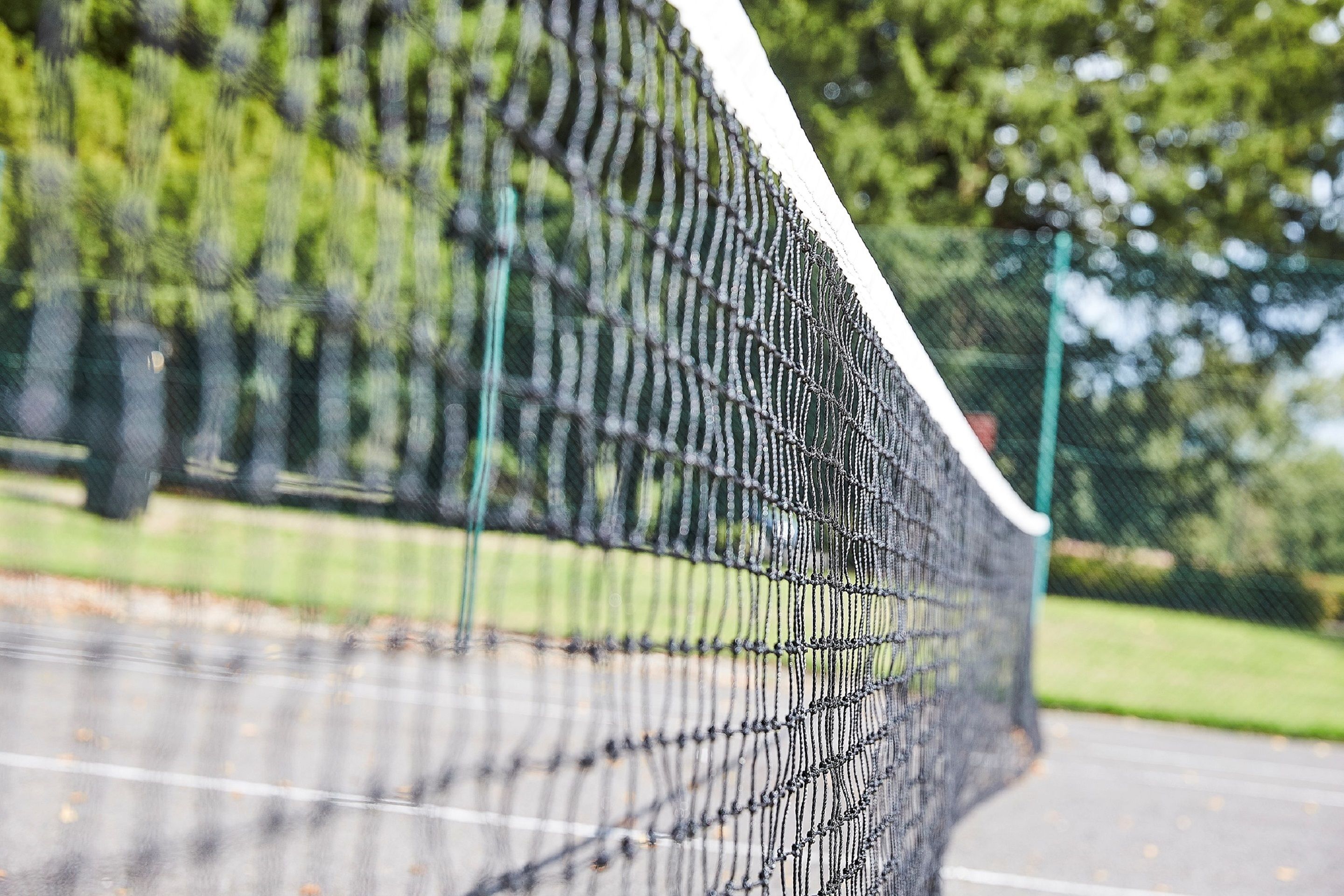 Lähikuva tenniskentän verkosta, taustalla epätarkkoja puita ja ruohikkoalue.