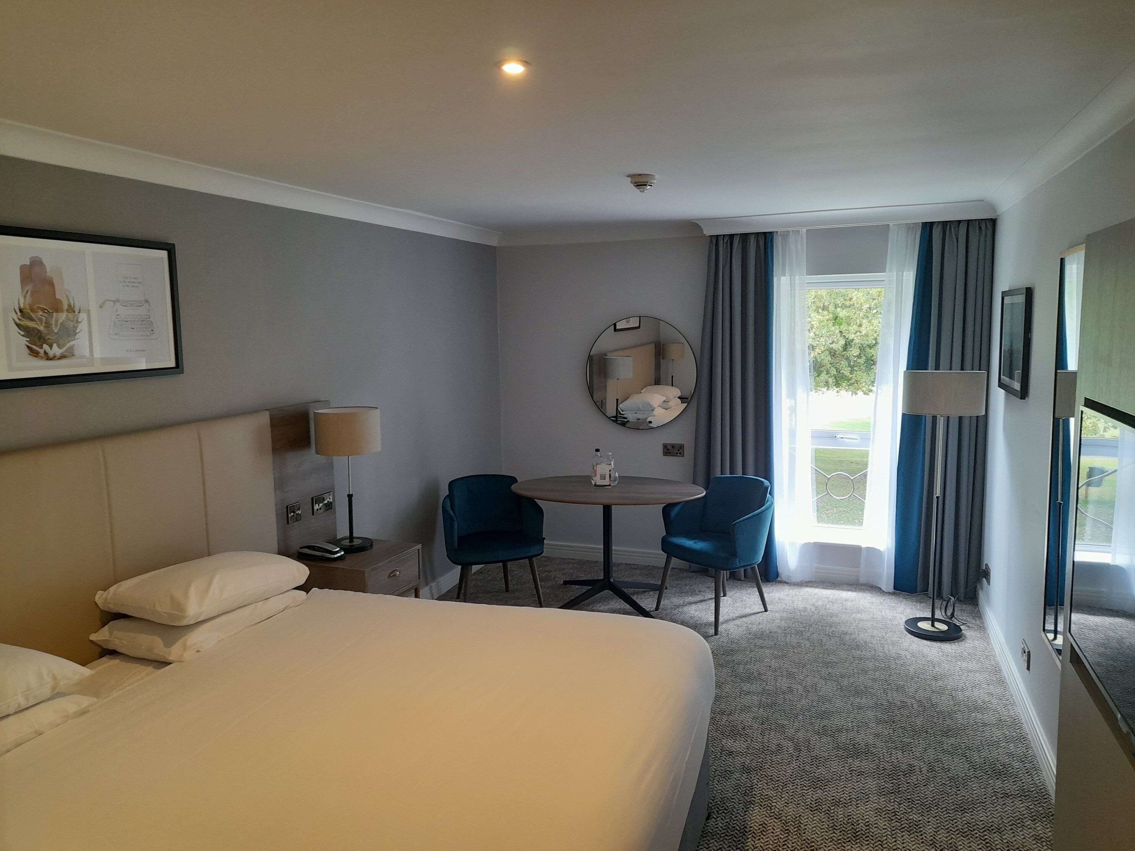 A hotel room with a bed, a desk with a chair, a round table, a mirror, and a window with curtains.