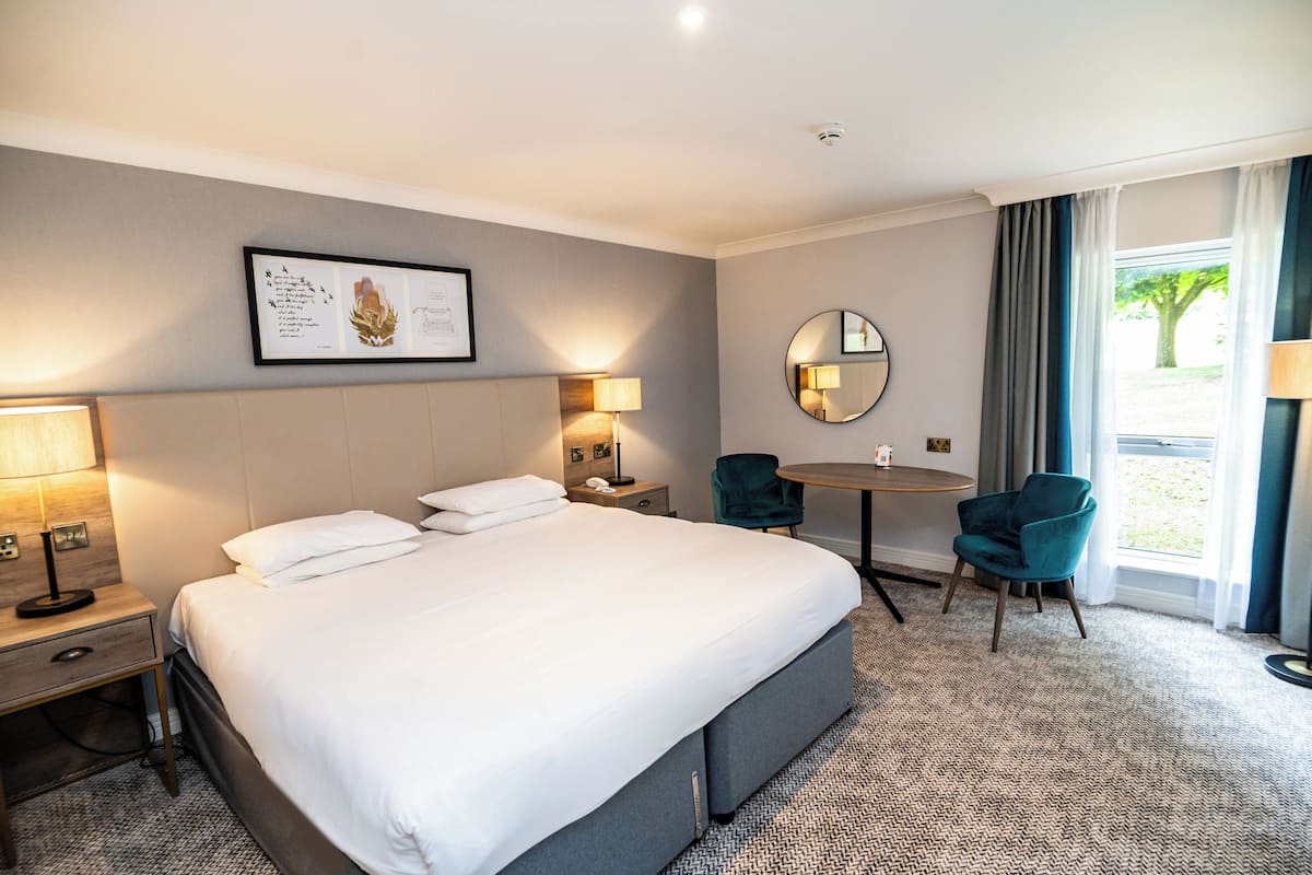 A hotel room with a large bed, bedside lamps, a round table, a chair, and a window with curtains.