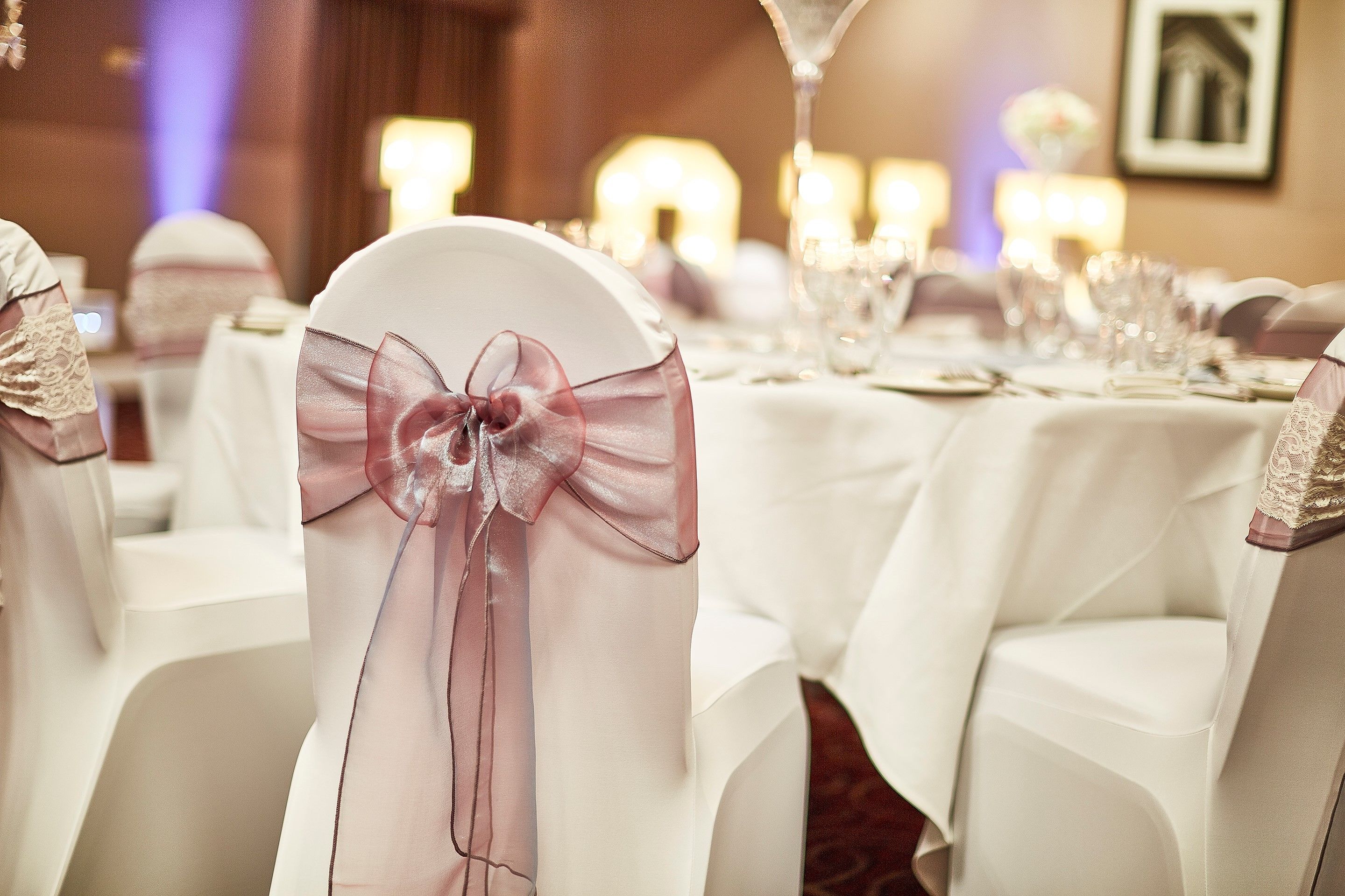 A banquet hall with round tables covered in white tablecloths, each chair adorned with a pink ribbon.