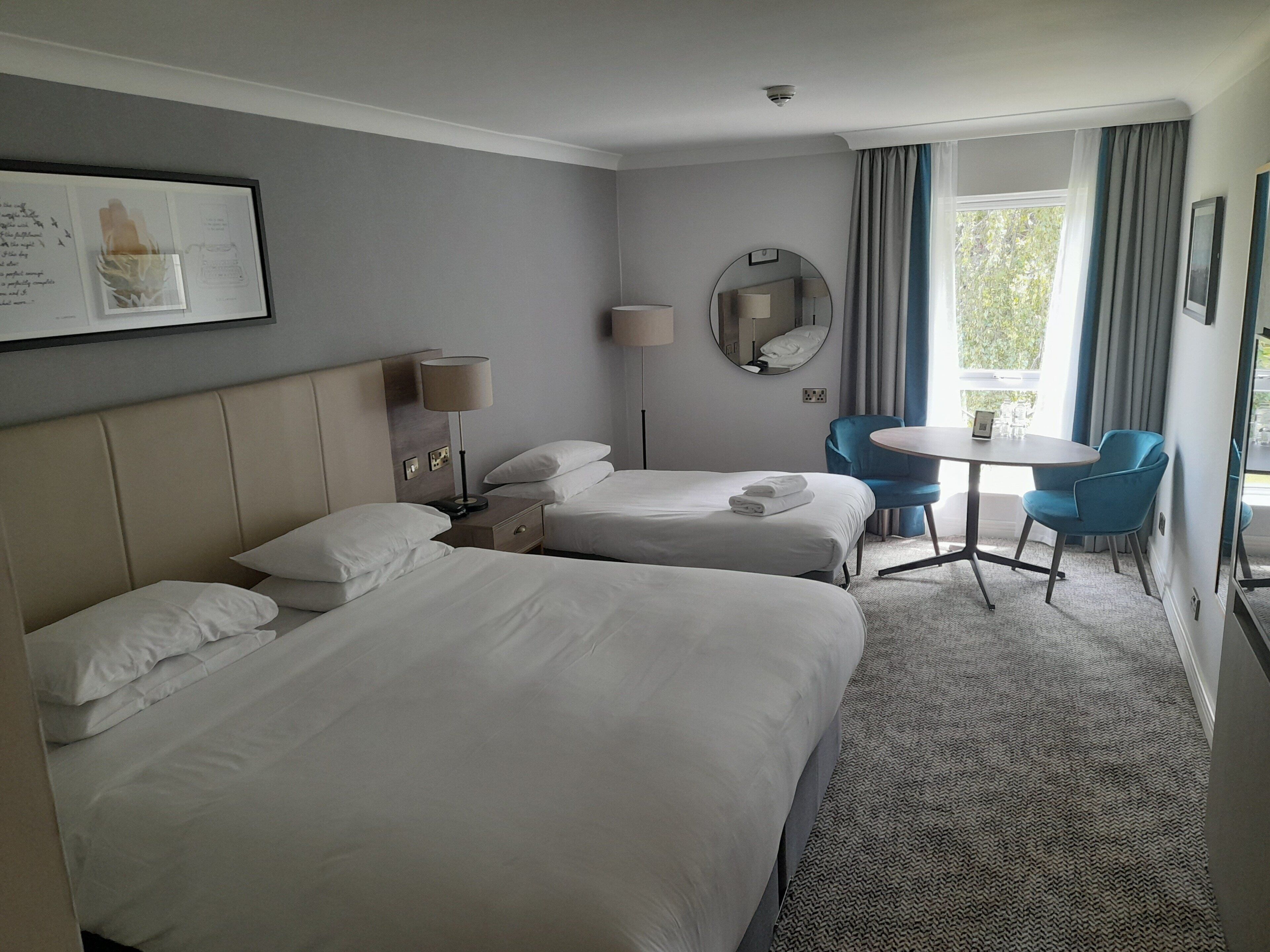 Habitación de hotel con dos camas, un escritorio, una silla, una mesa redonda, un espejo y una ventana con cortinas.