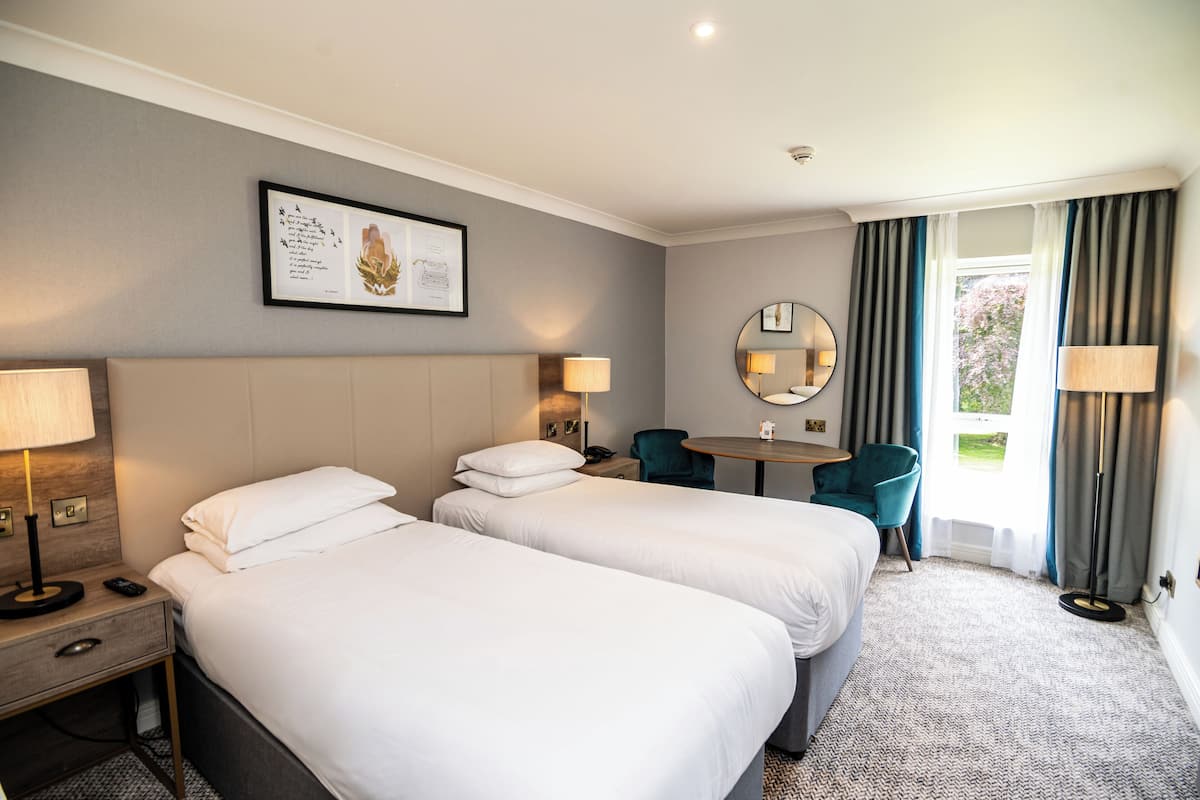 A hotel room with two beds, a round table, a chair, a desk, a lamp, a mirror, and a window with curtains.