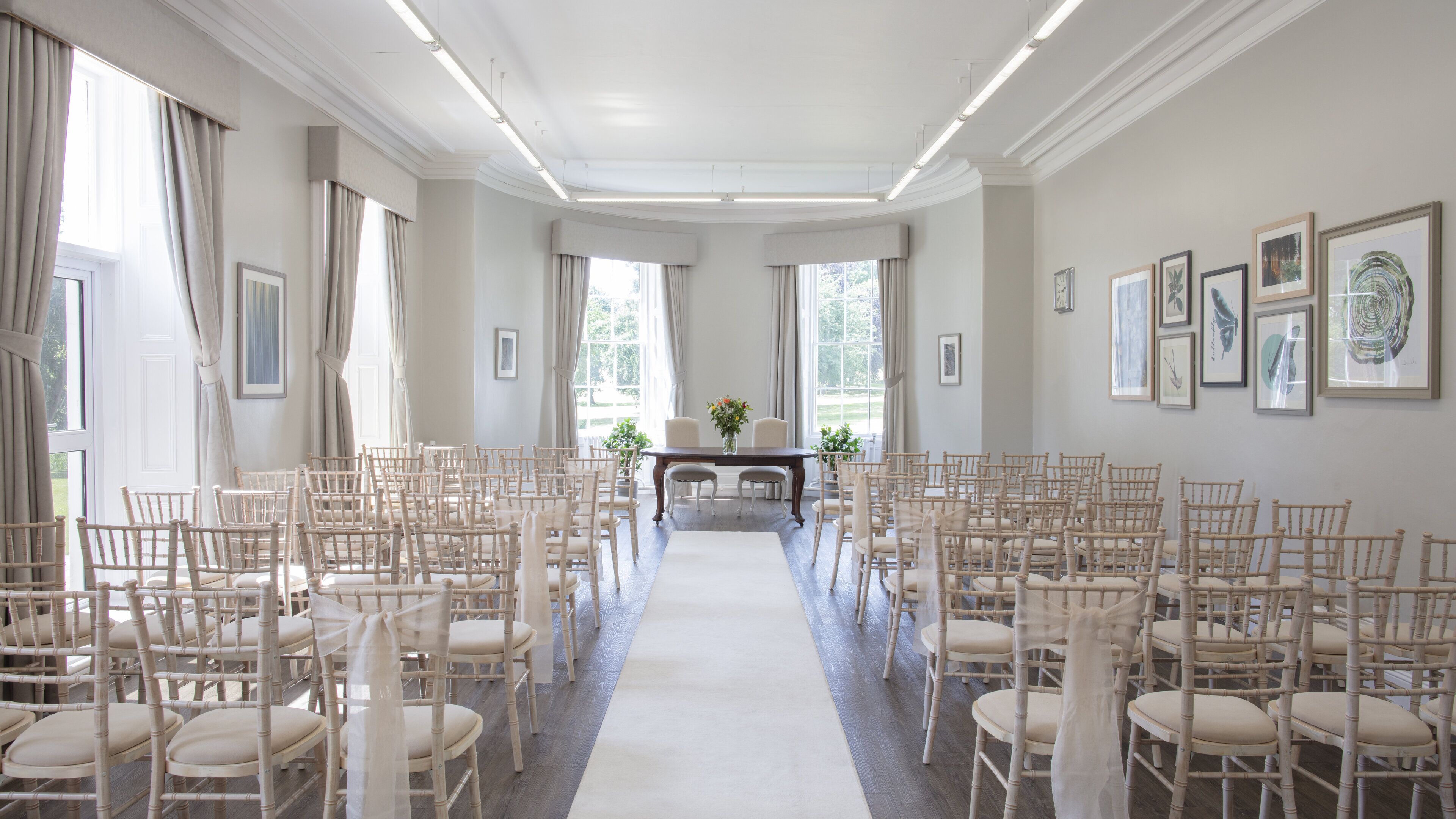 A formal room with rows of chairs arranged for an event, a central table with a vase of flowers, and framed artwork on the walls.
