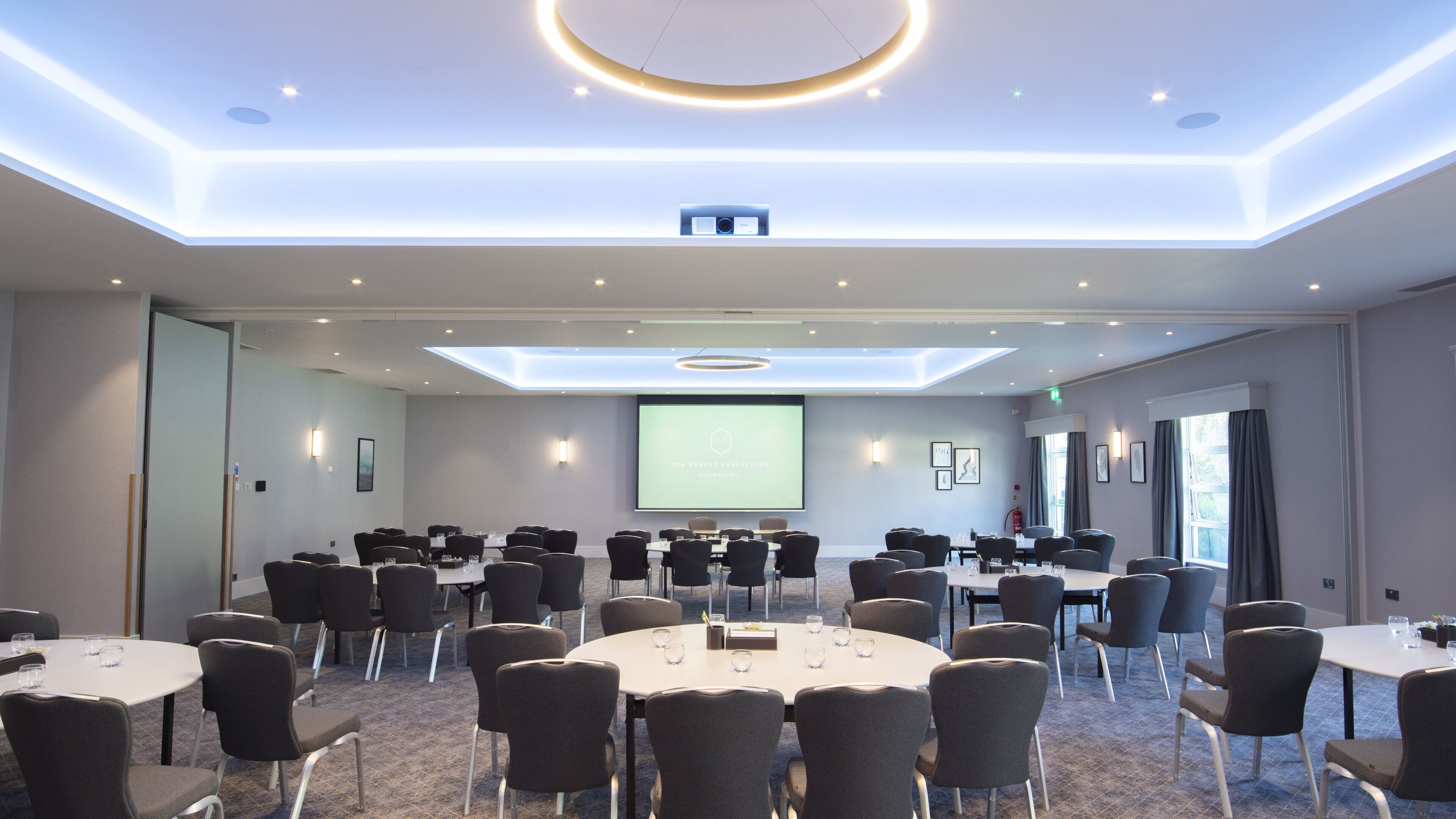 A conference room with multiple tables and chairs, a large screen at the front, and a skylight ceiling.