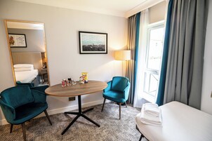 A hotel room with a bed, a round table with chairs, a lamp, a mirror, and a window with curtains.
