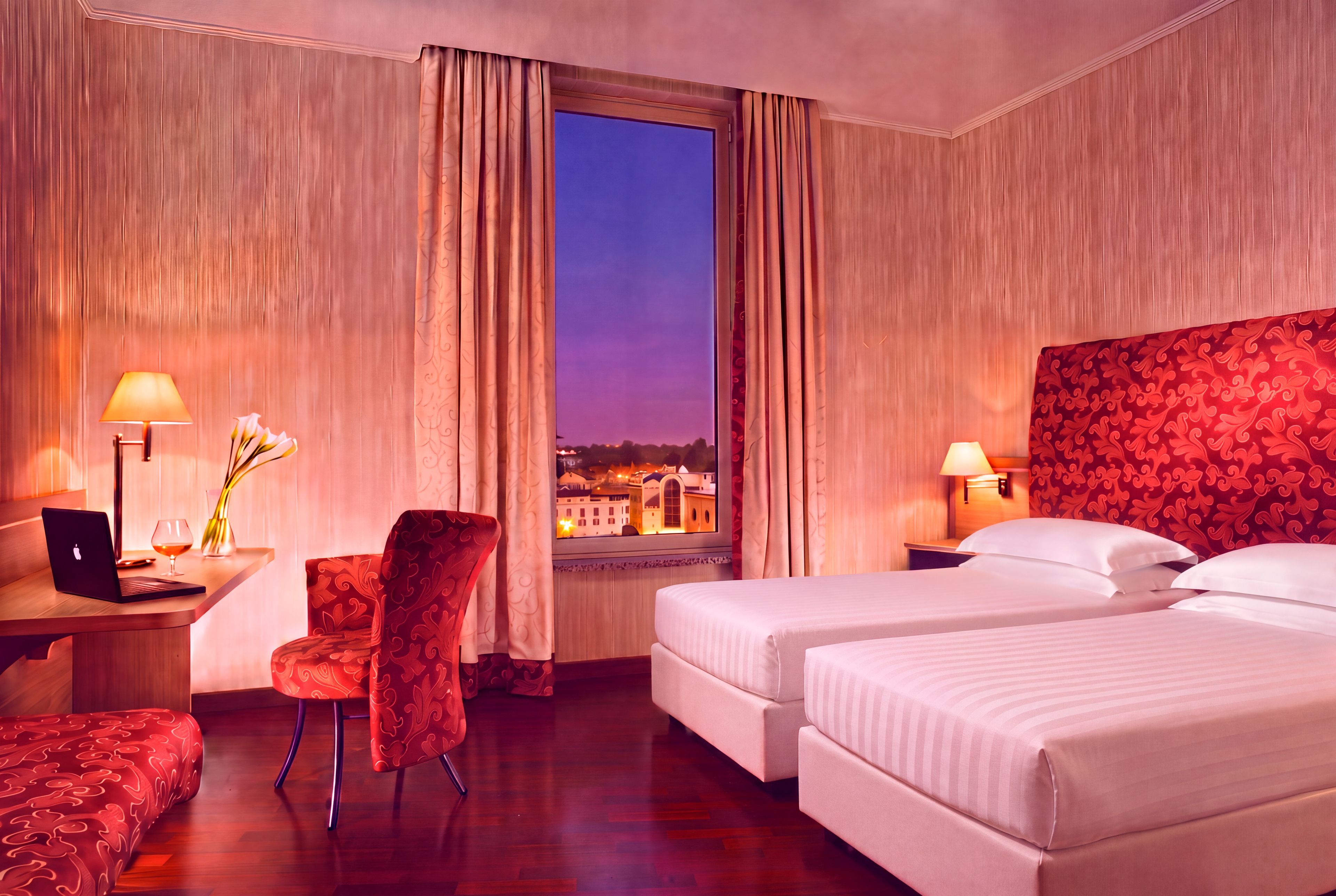 Une chambre d’hôtel équipée d’un bureau, d’une chaise, de deux lits et d’une fenêtre donnant sur des immeubles.