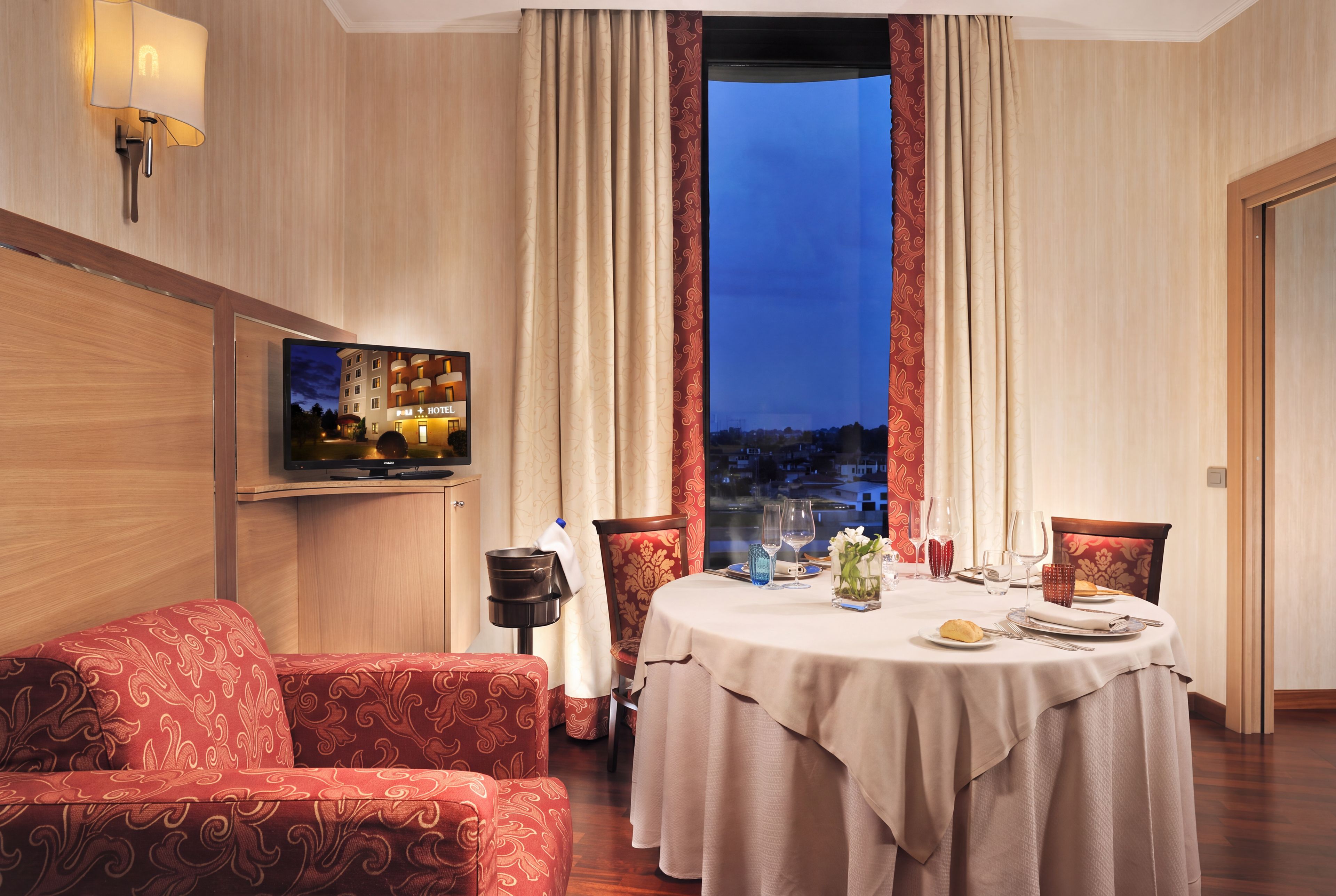 Ein Hotelzimmer mit Esstisch für zwei Personen, einer Couch, einem Fernseher und Blick auf die Stadtlandschaft.