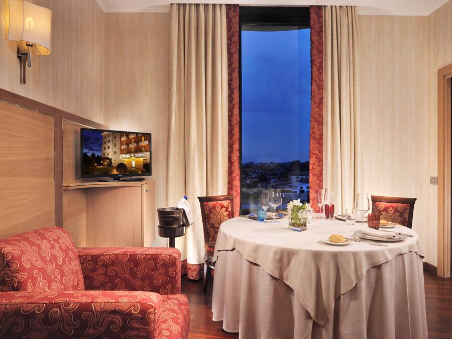 A hotel room with a dining table set for two, a sofa, a television, and a view of the cityscape.