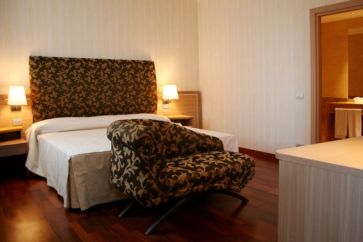 Habitación de hotel con cama, dos lámparas de noche, un sillón estampado y suelo de madera.