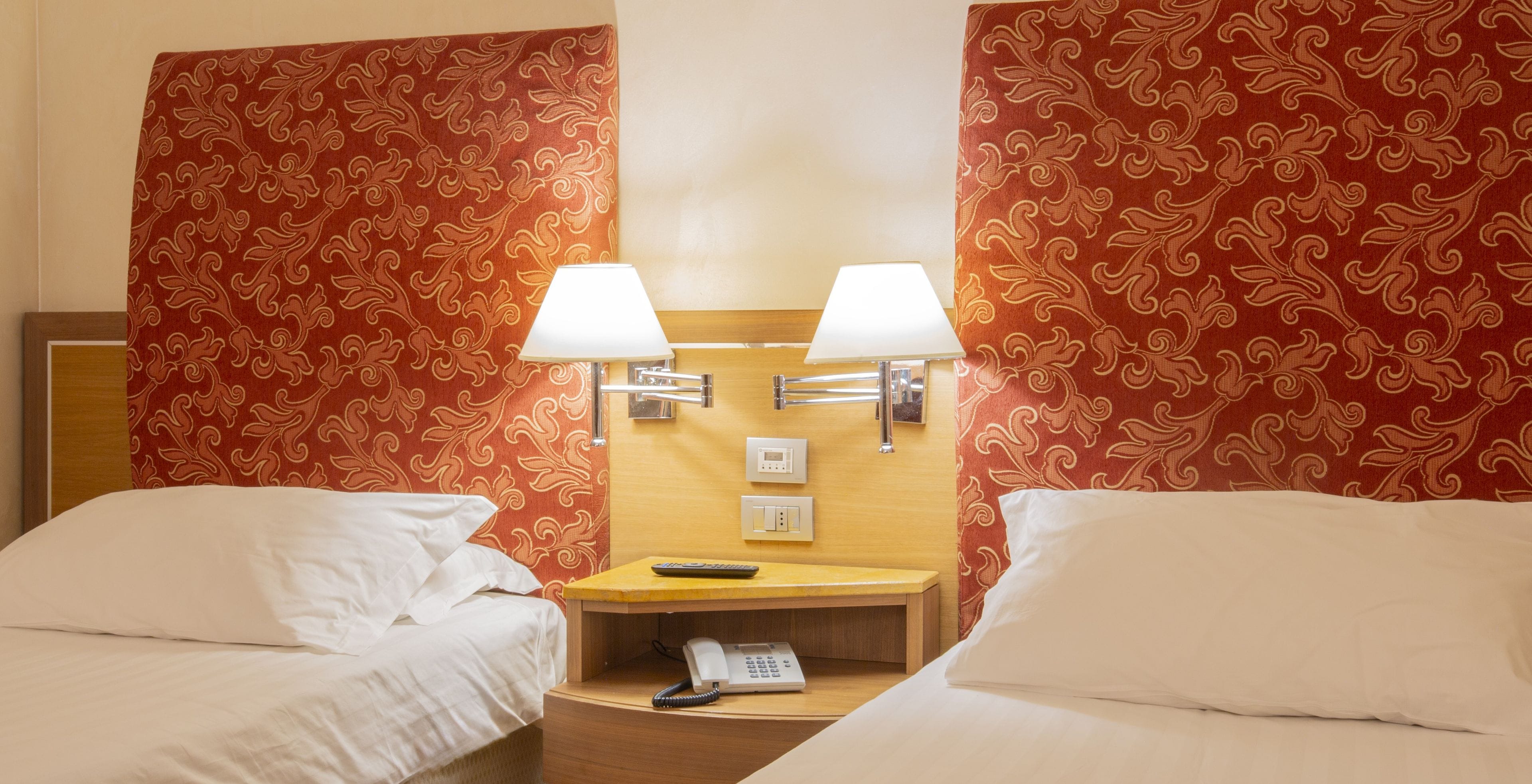 A hotel room with two beds, a bedside table with a lamp, a telephone, and a wall-mounted light switch.
