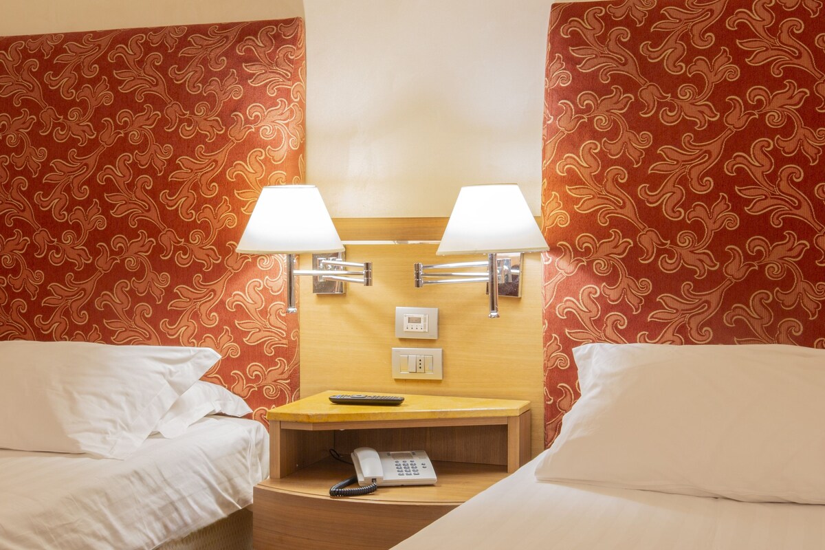 A hotel room with two beds, a bedside table with a lamp, a telephone, and a wall-mounted light switch.
