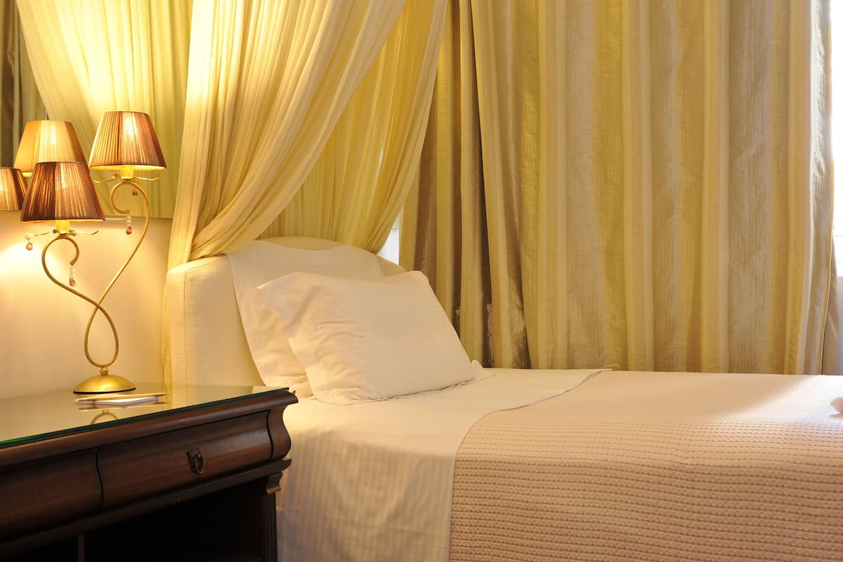 A neatly made bed with white linens and a single white pillow, a bedside table with a lamp, and sheer curtains in a hotel room.