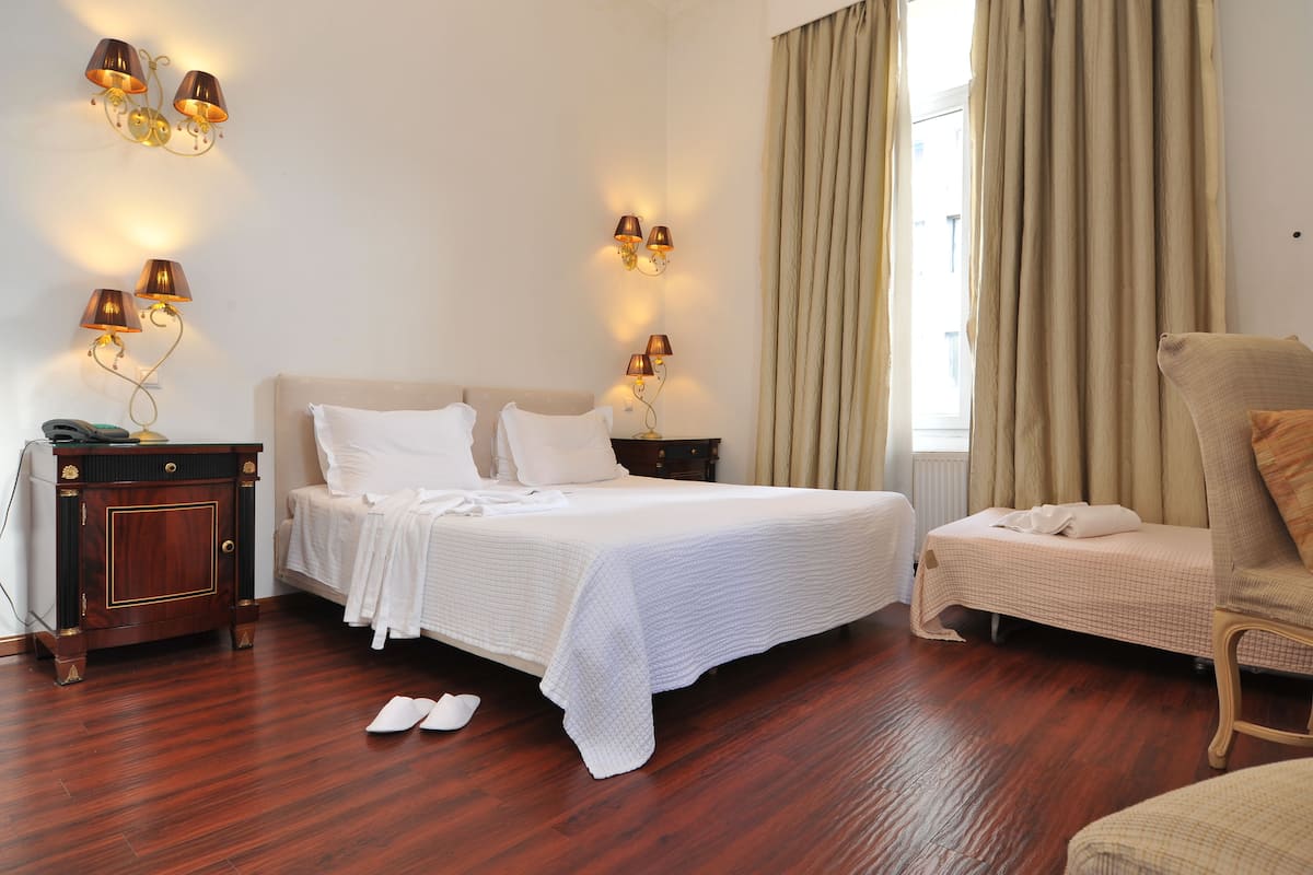 A neatly made bed with white linens, a wooden nightstand with a lamp, and a chair in a room with wooden flooring and beige curtains.