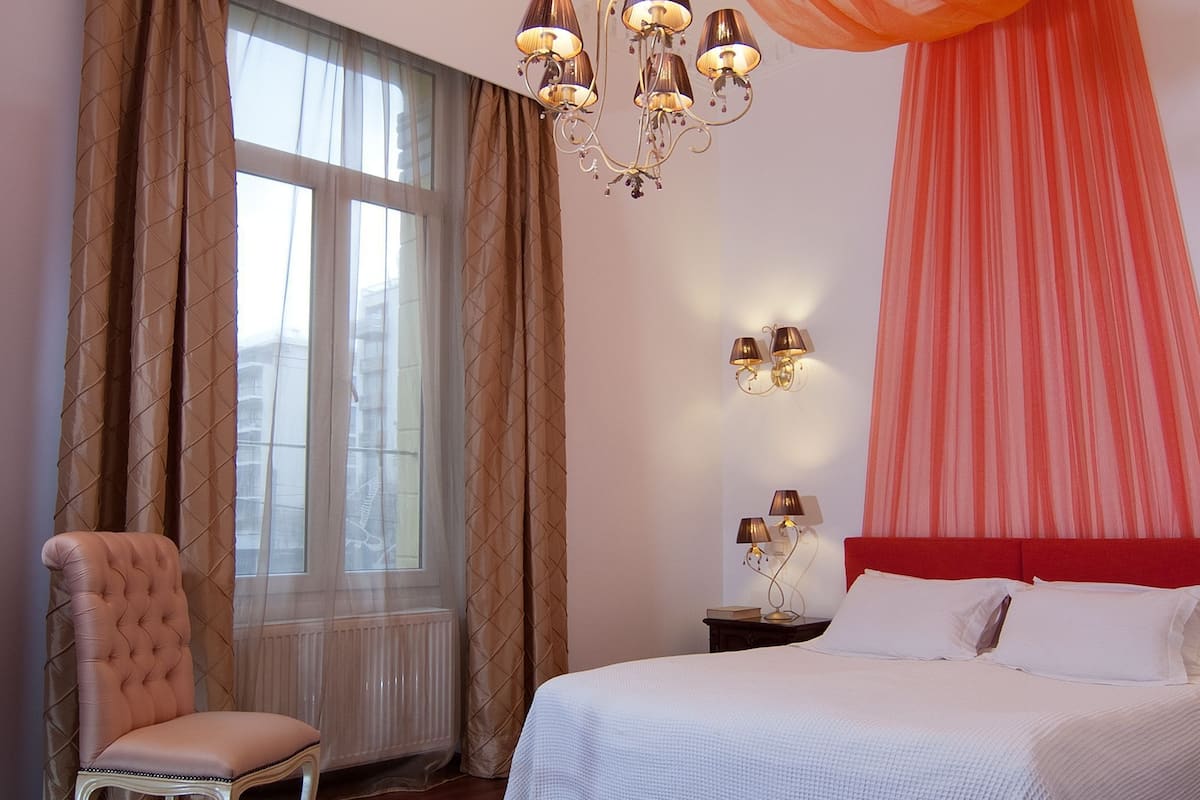 A bedroom with a bed, a chair, a window with curtains, and a chandelier.