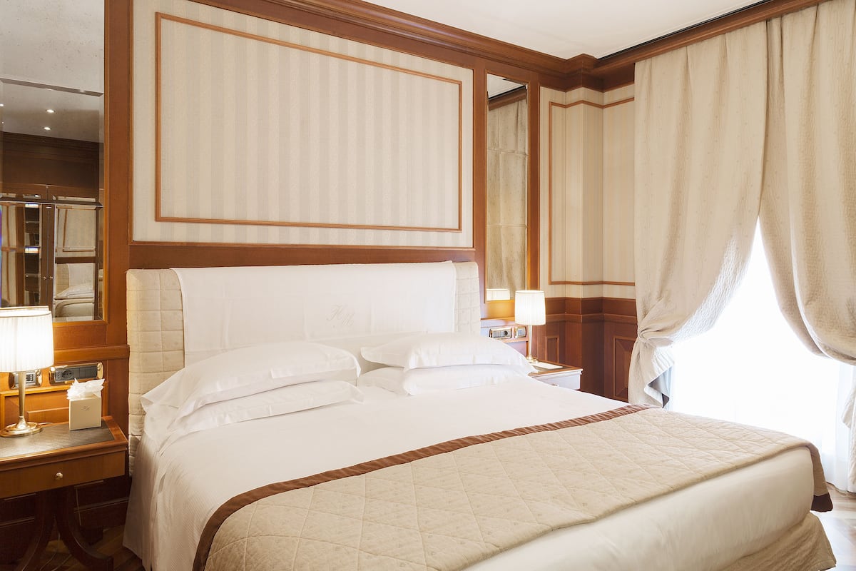 Habitación de hotel con una cama grande, una mesita de noche con lámpara, un espejo y una ventana con cortinas.