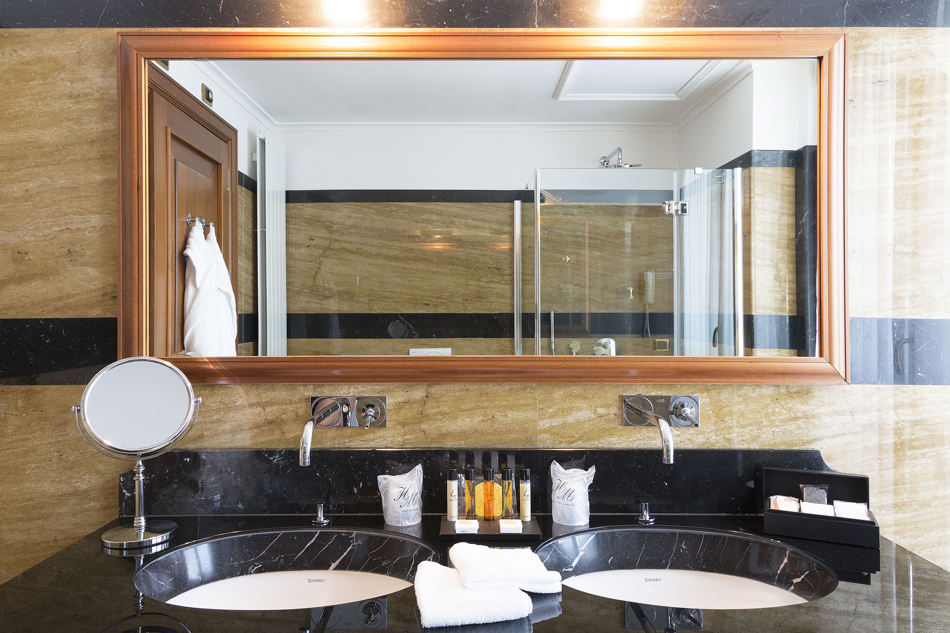 A bathroom with two sinks, a large mirror, and a shower visible in the reflection.