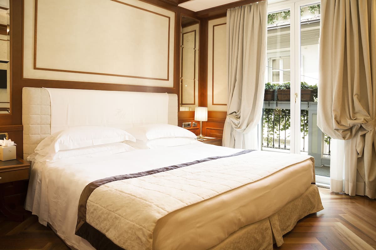Una cama bien hecha con una colcha blanca y marrón, un cabecero de madera, una mesita de noche con una lámpara, y vistas al exterior a través de una puerta corrediza de vidrio.